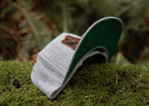 Hiking Boot, Flat Bill Hat resting on moss with a faux leather patch, designed for outdoor adventures.