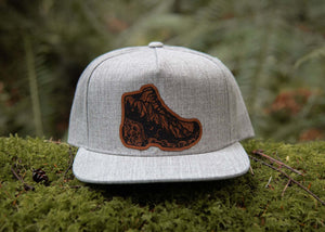Hiking Boot, Flat Bill Hat on moss, featuring faux leather patch and adjustable snapback, perfect for outdoor adventures.