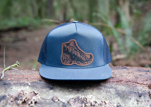 Hiking Boot, Flat Bill Hat displayed on a log in a forest, featuring a faux leather patch and mountain design.