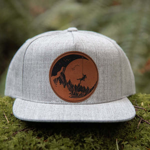 Moon and Climber, Flat Bill Hat showcased on moss background, featuring faux leather patch and adjustable snapback.
