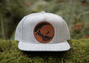 Moon and Climber Flat Bill Hat showcased on moss background, featuring faux leather patch and adjustable snapback.