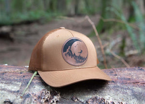 Moon and Climber, Trucker Hat on a log in the forest, showcasing a durable design by a Canadian artist.