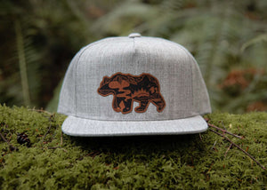 Mountain Bear Flat Bill Hat displayed on moss with a durable faux leather patch, ideal for outdoor adventures.
