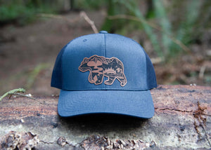 Mountain Bear Trucker Hat on a log in nature, designed for outdoor adventures, by a Canadian artist.