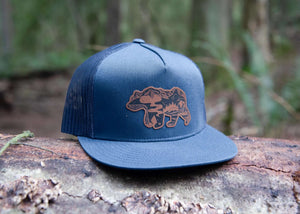 Mountain Bear Flat Bill Hat on a log, featuring a durable faux leather patch and breathable mesh back.