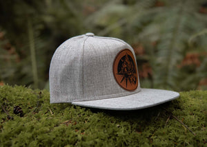 Sea to Sky flat bill hat on mossy ground, featuring faux leather patch and perfect for outdoor adventures.