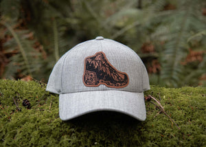 Hiking Boot, Trucker Hat with durable faux leather patch on a mossy forest floor.