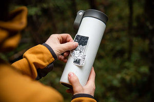 Person applying an Alberta Sticker on a white travel mug in a forest setting, showcasing outdoor sticker design.