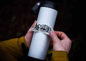 Person applying Bear and Mountain Sticker to a water bottle, showcasing a black and white outdoor sticker design.