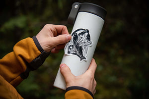 Person applying a Bear & Salmon Sticker to a travel mug, showcasing outdoor sticker design by a Canadian artist.