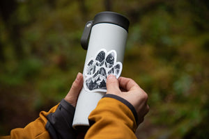 Dog Paw Sticker applied on a travel mug outdoors, featuring a black and white original illustration by a Canadian artist.