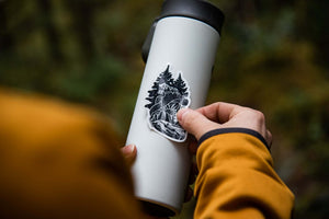 Person applying a Forest Heart Sticker on a white travel mug in the outdoors.
