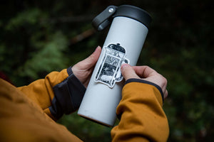 Person applying a French Press Sticker to a stainless steel water bottle outdoors, showcasing the black and white vinyl design.