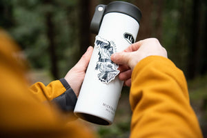 Jasper National Park Sticker being applied to a white thermos in the outdoors, showcasing an original illustration.