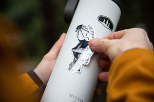 Person applying a Kootenay National Park Sticker to a white water bottle outdoors, showcasing the original illustration.