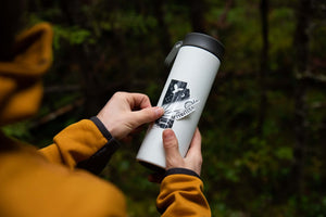 Manitoba Sticker on a white travel mug held by a person in outdoor clothing, showcasing original black and white illustration.