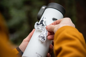 Person applying Newfoundland and Labrador sticker on a white travel mug outdoors, showcasing durable vinyl design.