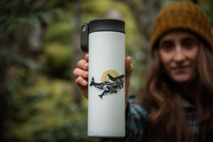 Person holding a travel mug featuring the Orca Baby Sticker, a black and white vinyl outdoor sticker.