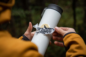 Orca Baby Sticker on a travel mug, showcasing black and white original illustration. Durable outdoor sticker made in Canada.
