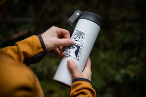 Person applying Saskatchewan Sticker to a tumbler outdoors, showcasing an outdoor sticker design by a Canadian artist.