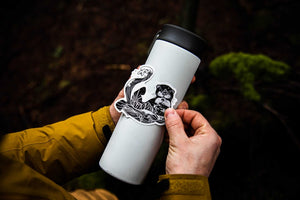 A person applying a Sea Otter Sticker on a white tumbler outdoors, showcasing an original illustration by a Canadian artist.