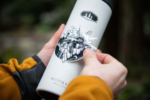 Person applying the Sea to Sky Sticker to a water bottle outdoors, showcasing the black and white original illustration.