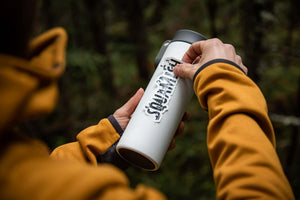 Person applying a Squamish Sticker on a white insulated tumbler in a natural outdoor setting.