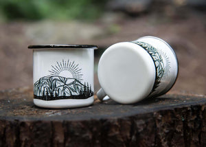The Stawamus Chief Enamel Mug displayed outdoors, showcasing Canadian artist's original illustration on enamel drinkware.