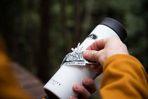 Three Sisters Mountain Sticker applied to a travel mug in a natural outdoor setting, showcasing a black and white original illustration.
