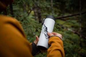 Person applying a Vancouver Island Sticker on a travel mug in a forest, showcasing outdoor sticker and Canadian art.